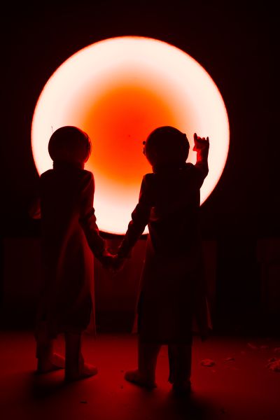 Nina Noé Stehlin und Henriette Nagel in ihren Rollen stehen mit dem Rücken zum Publikum. Sie tragen Weltraumhelme und weiße Kittel. Sie blicken auf ein großes kreisförmiges helles Licht mit einem orangefarbenen Kern in der Mitte.